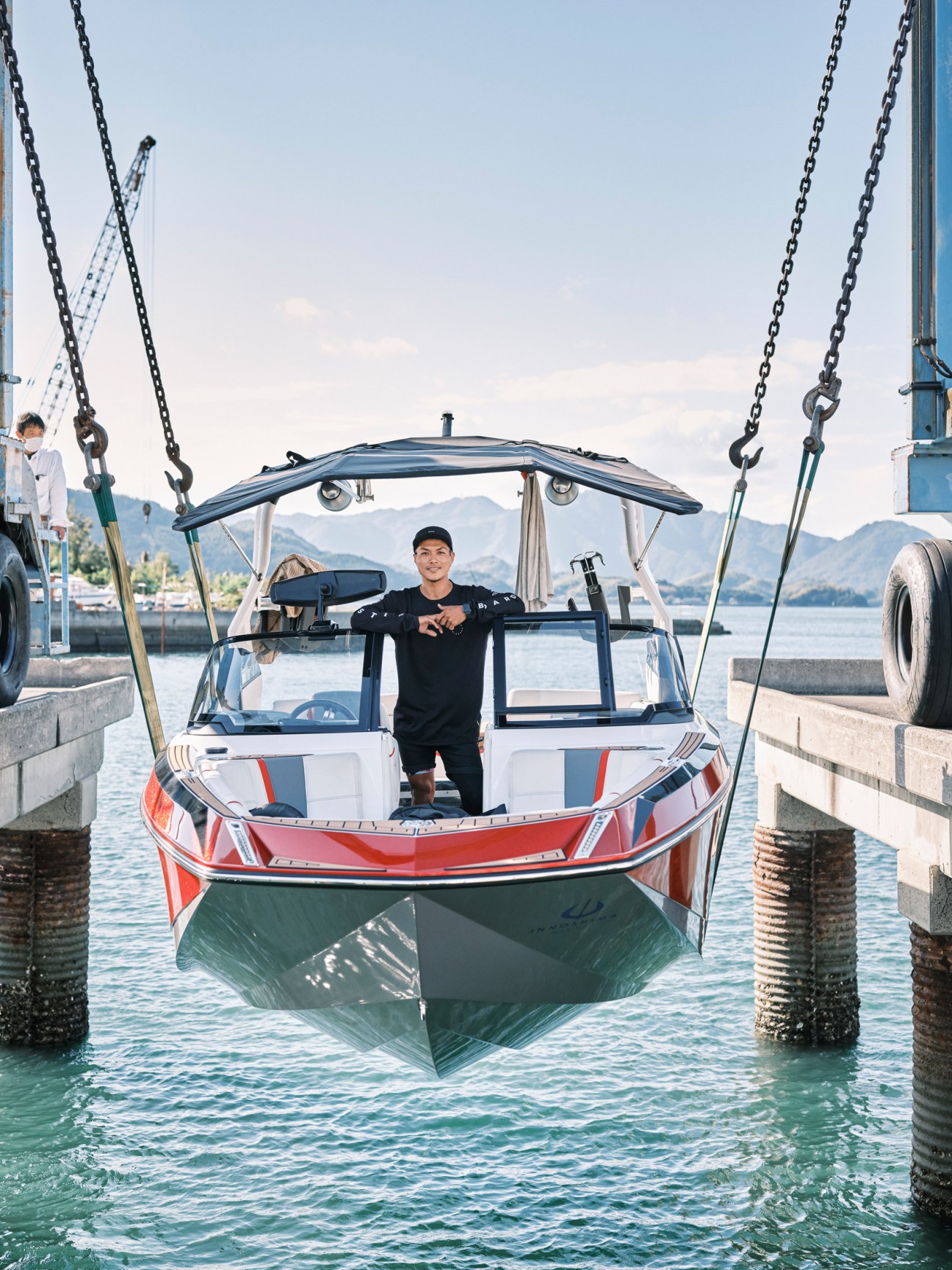 Man in black shirt stands in red and green speedboat suspended by chains over water with mountains in background.