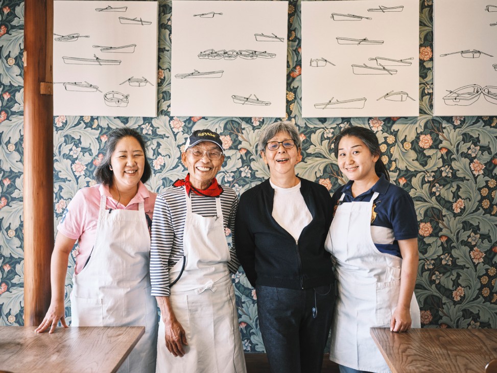 Four people wearing aprons standing together smiling in front of floral wallpaper and boat drawings