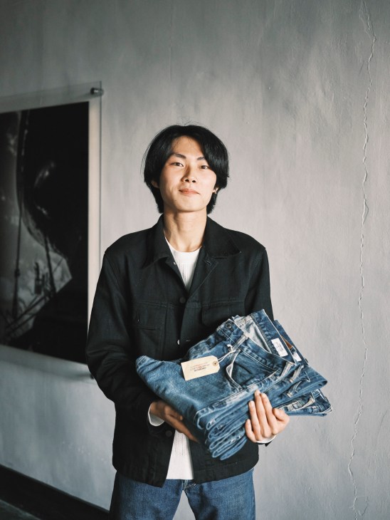 Young man in black jacket holding folded denim jeans against white wall