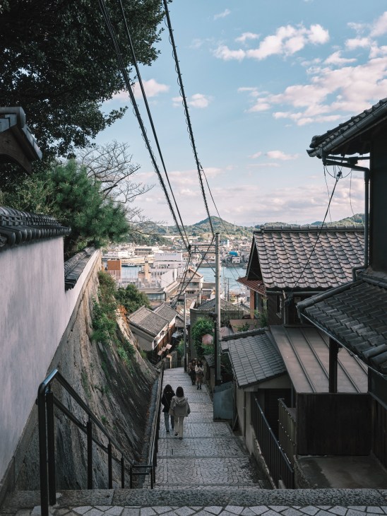 Narrow hillside alley lined with traditional Japanese buildings overlooking Onomichi's harbor and waterfront.
