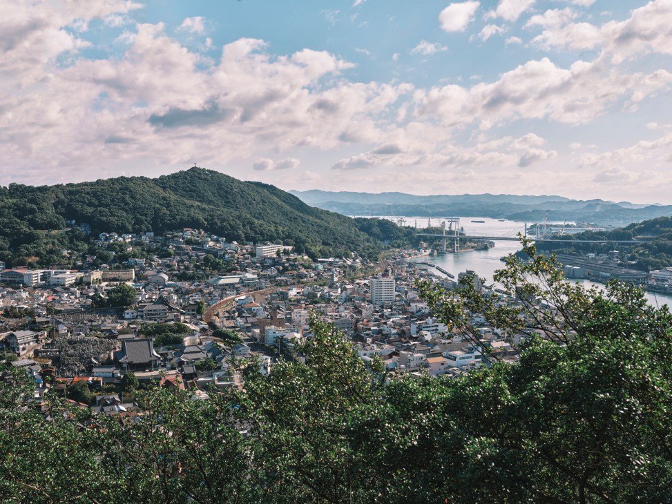Onomichi cityscape viewed from hillside overlooking residential buildings, forested slopes and the Seto Inland Sea with shipyards.