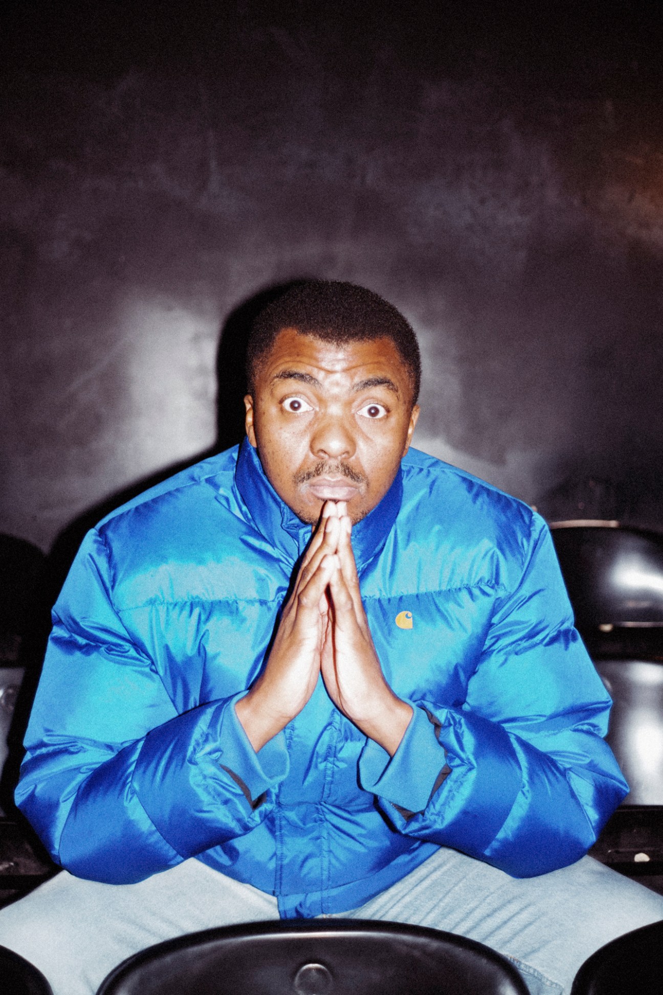 Loyiso Gola in a blue puffy jacket making a prayer gesture with wide-eyed expression