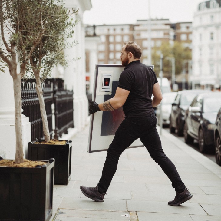 Person in black clothing moving a large framed artwork or monitor on a street sidewalk