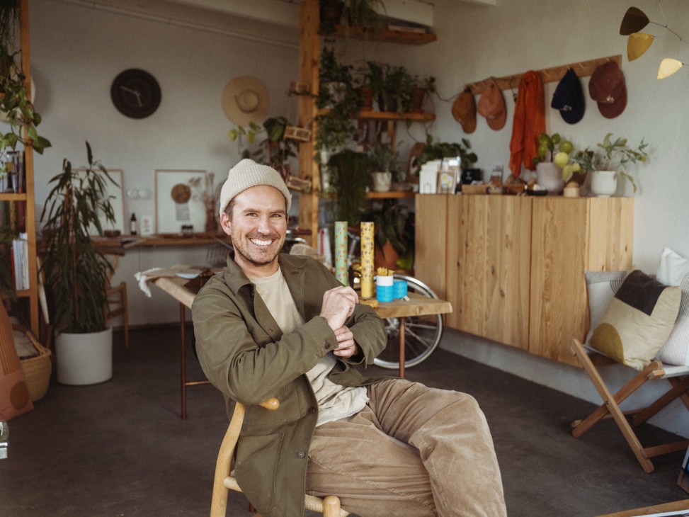 Man in beige knit hat and olive jacket sitting in modern interior with wooden furniture, plants, and hats on wall