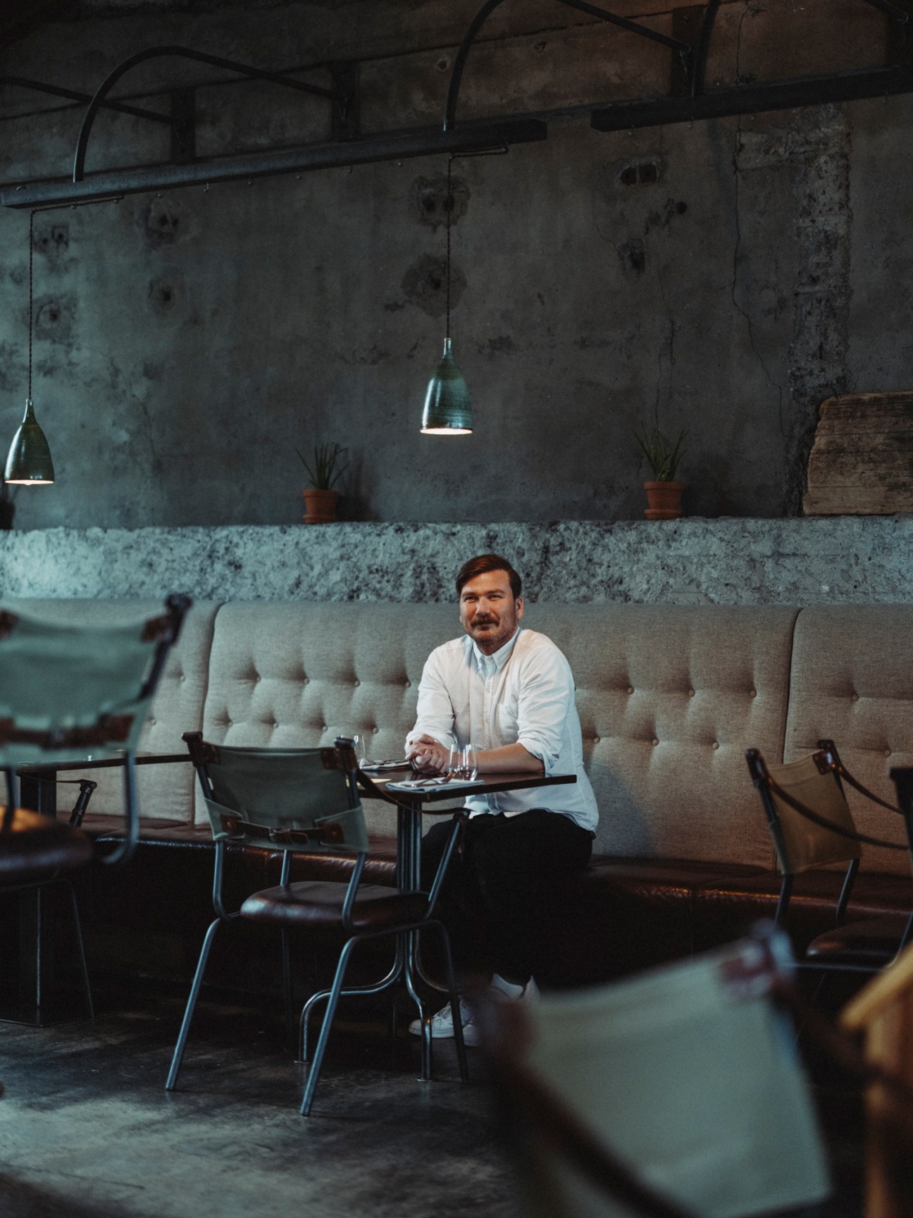 Man with mustache in white shirt sitting at table in industrial-style restaurant with concrete walls and pendant lighting