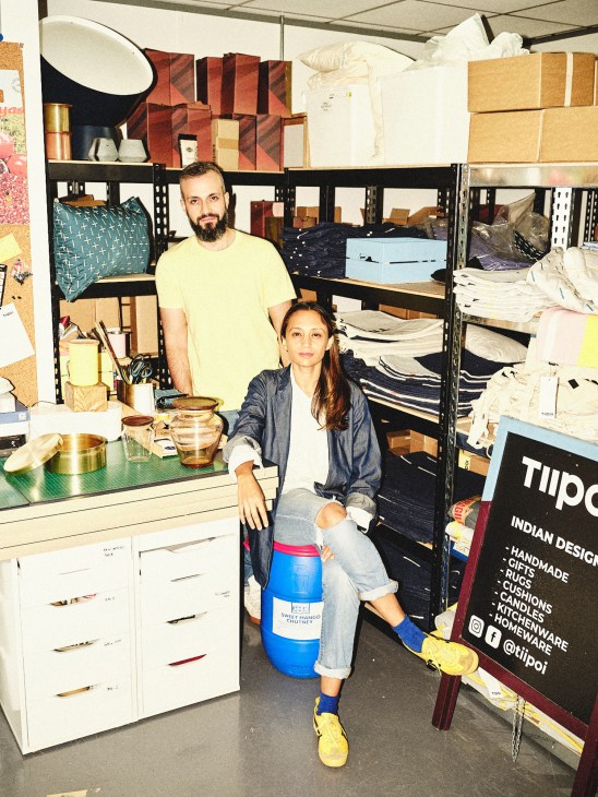 Two people posing in Tiipoi's studio workspace surrounded by shelves of crafted products and materials.