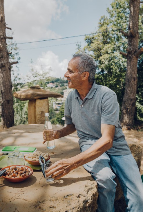 Charbel Bteish sits at a stone table in his mountain garden, holding a bottle while bowls of nuts sit before him.