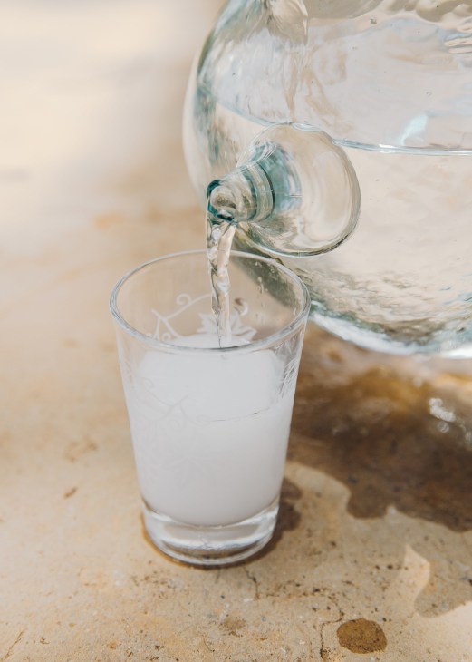 Homemade arak being poured from a glass jug into a small glass on a sandy surface.
