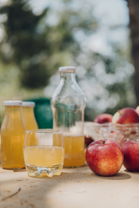 Fresh apple juice in glasses and bottles displayed with red apples in a garden setting.