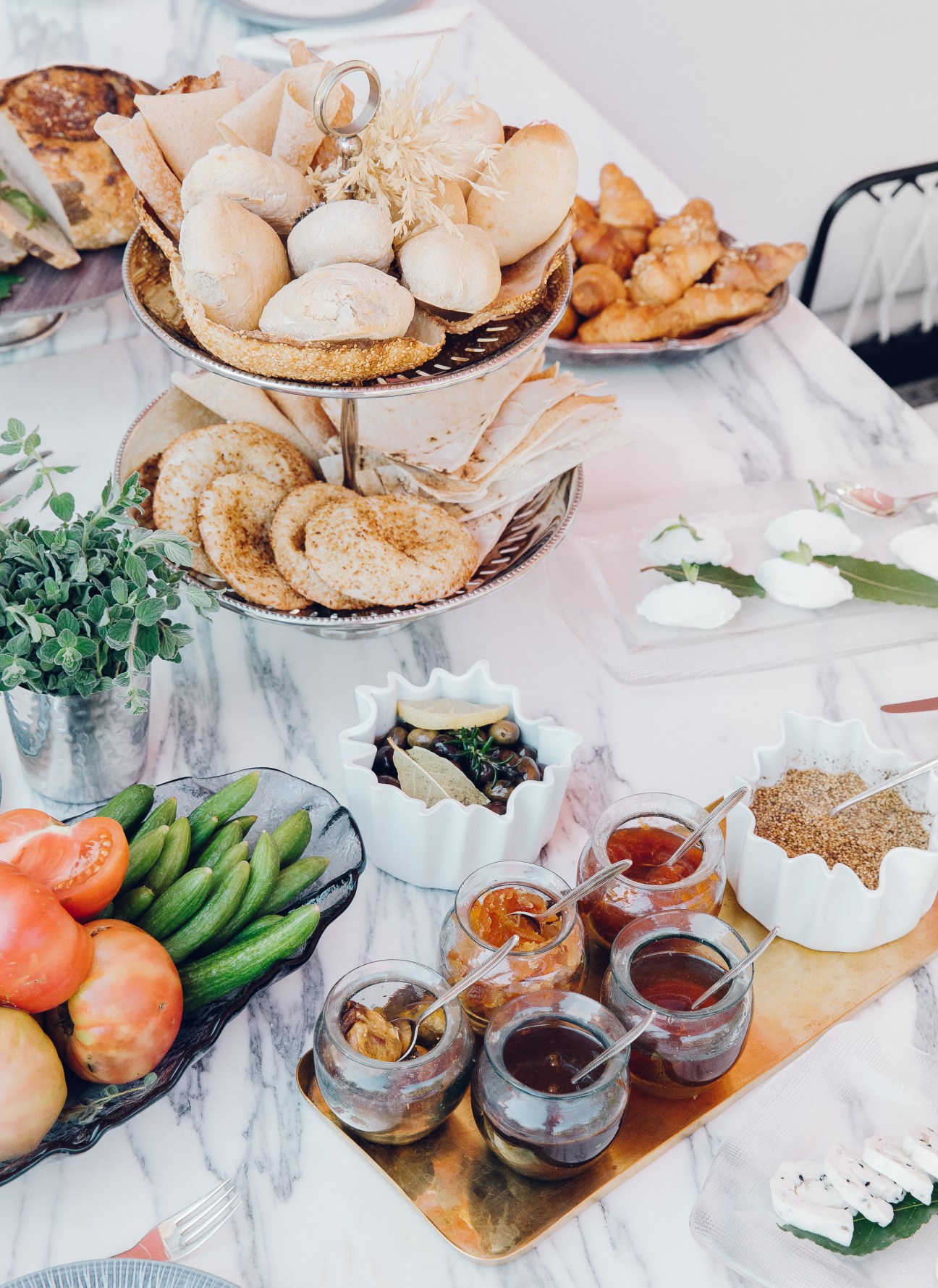A tiered breakfast display with pastries, vegetables, jams, and fresh herbs on marble surface.