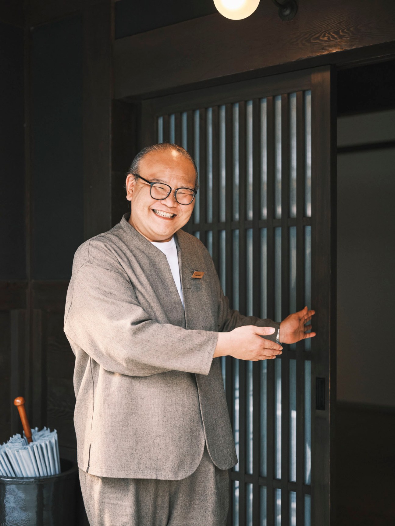 Seiji Murakami in grey suit and glasses smiling while gesturing welcomingly at traditional Japanese entrance