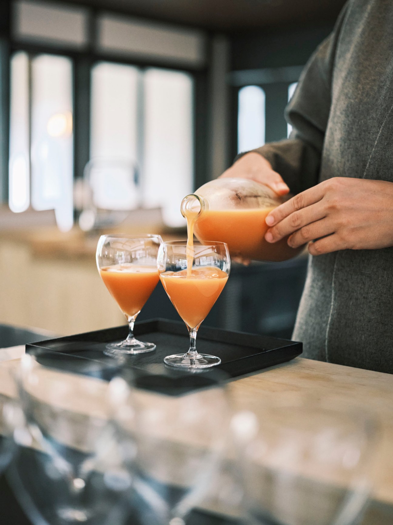 Person pouring orange citrus juice from glass pitcher into two elegant glasses on black tray