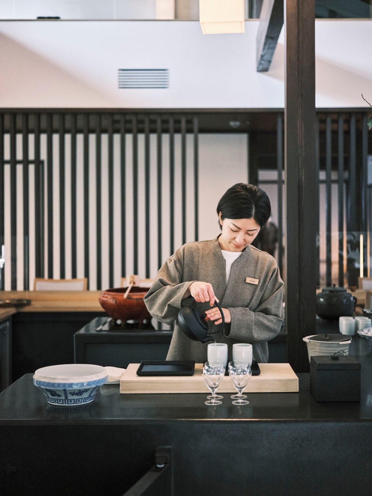 Woman in grey traditional Japanese clothing preparing green tea service with ceramic teapot and cups on wooden counter