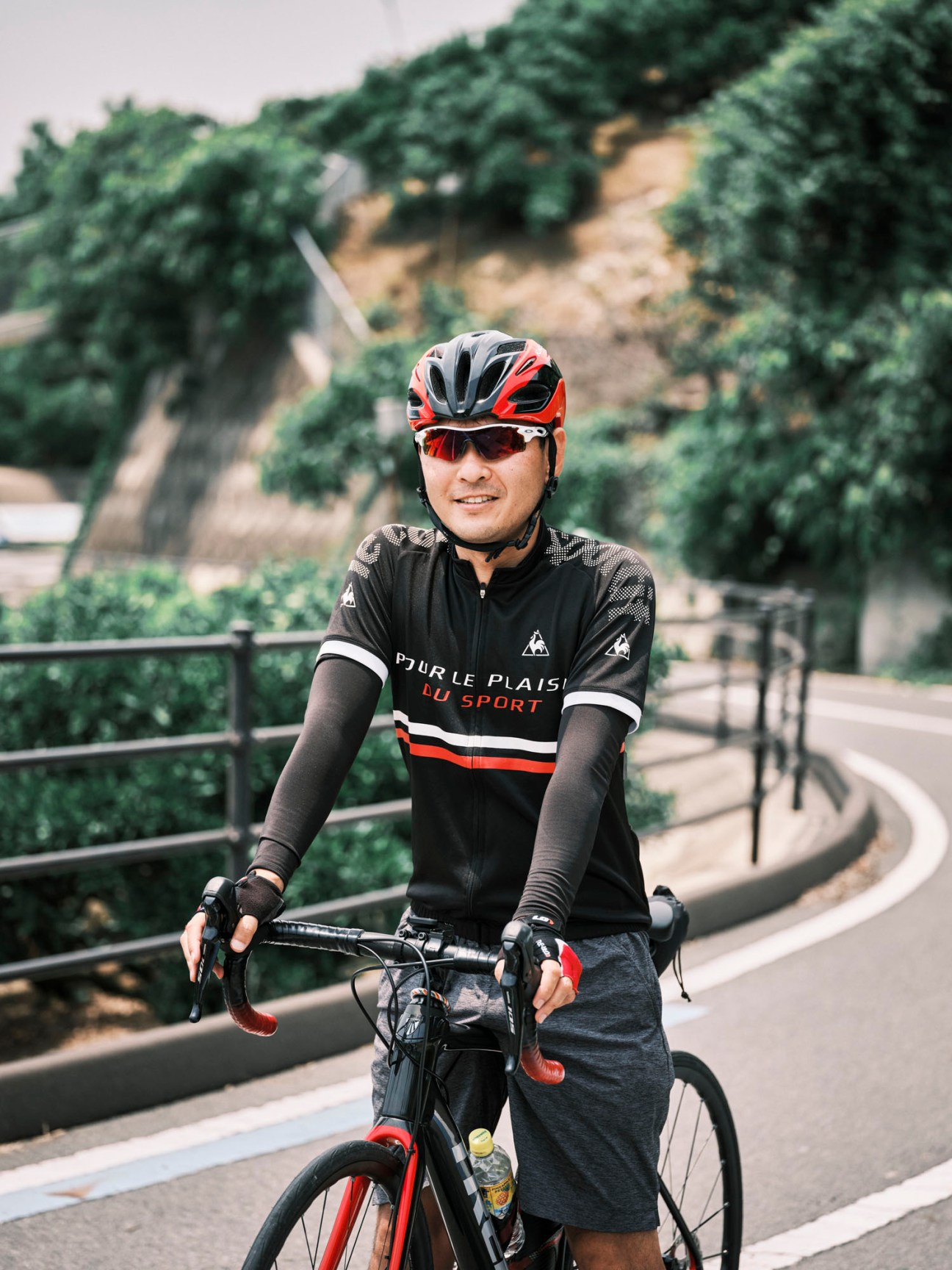 Cyclist in red and black helmet with Le Coq Sportif jersey standing with bicycle on curved path