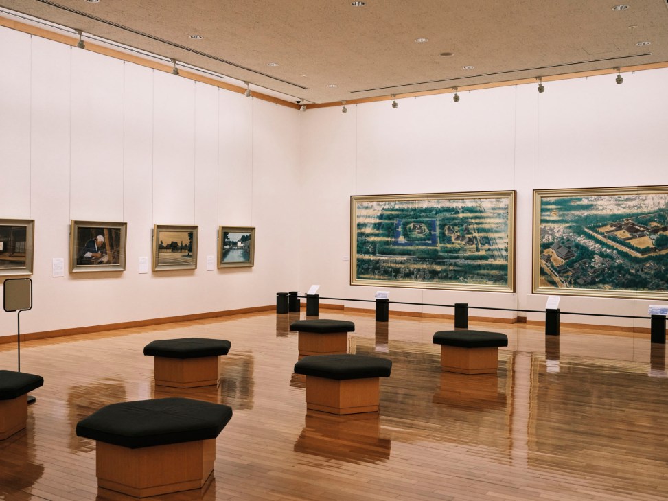 Museum gallery interior with paintings on white walls, wooden floors, and black cushioned benches