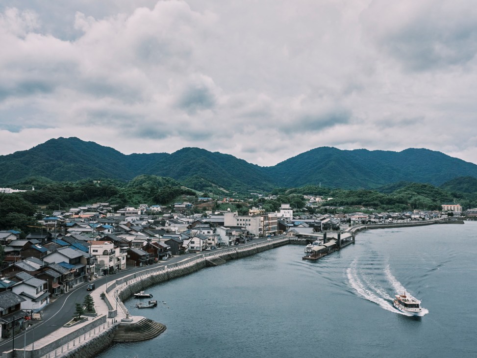 Aerial view of Setoda port town with traditional buildings along waterfront, forested mountains behind, and ferry boat in harbor