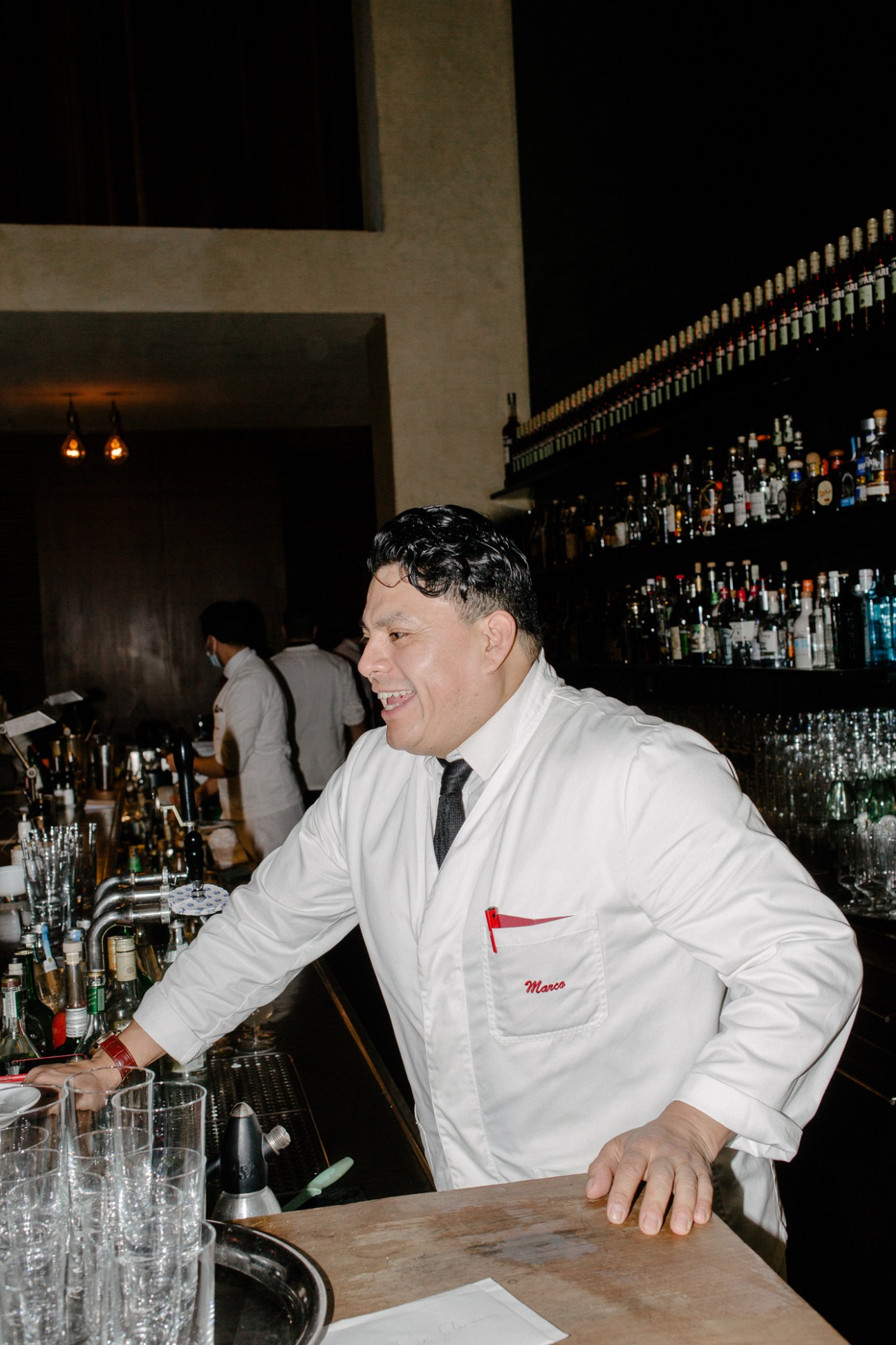 Smiling bartender in white uniform with name tag behind bar counter at Schumann's
