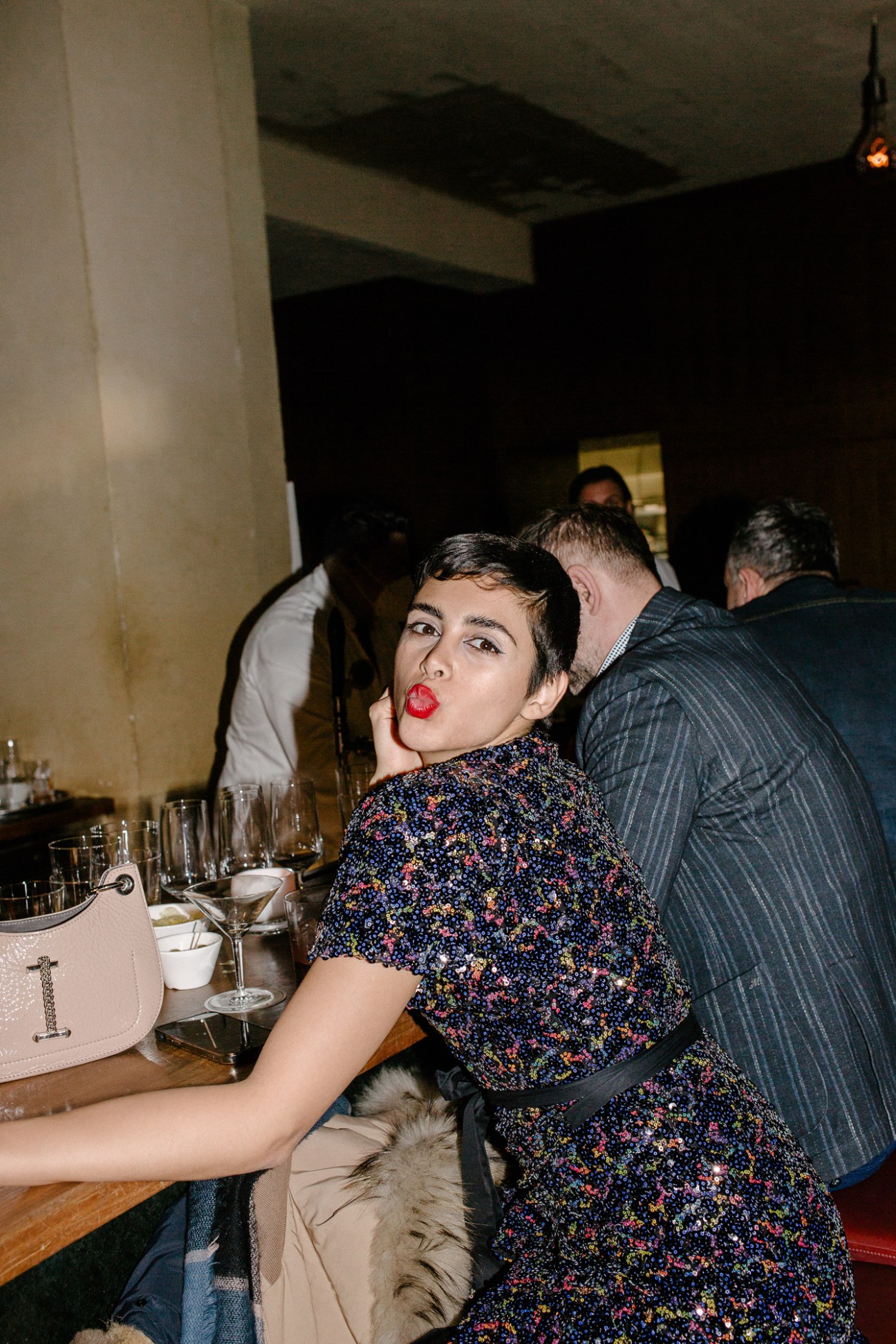 Woman with short dark hair and red lipstick making kissing gesture at camera in dimly lit bar setting