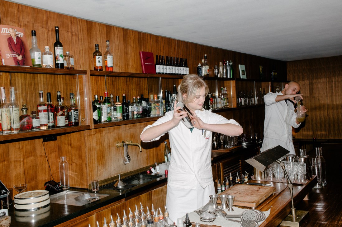 Two bartenders in white uniforms working behind a wood-paneled bar with extensive liquor bottles on shelves