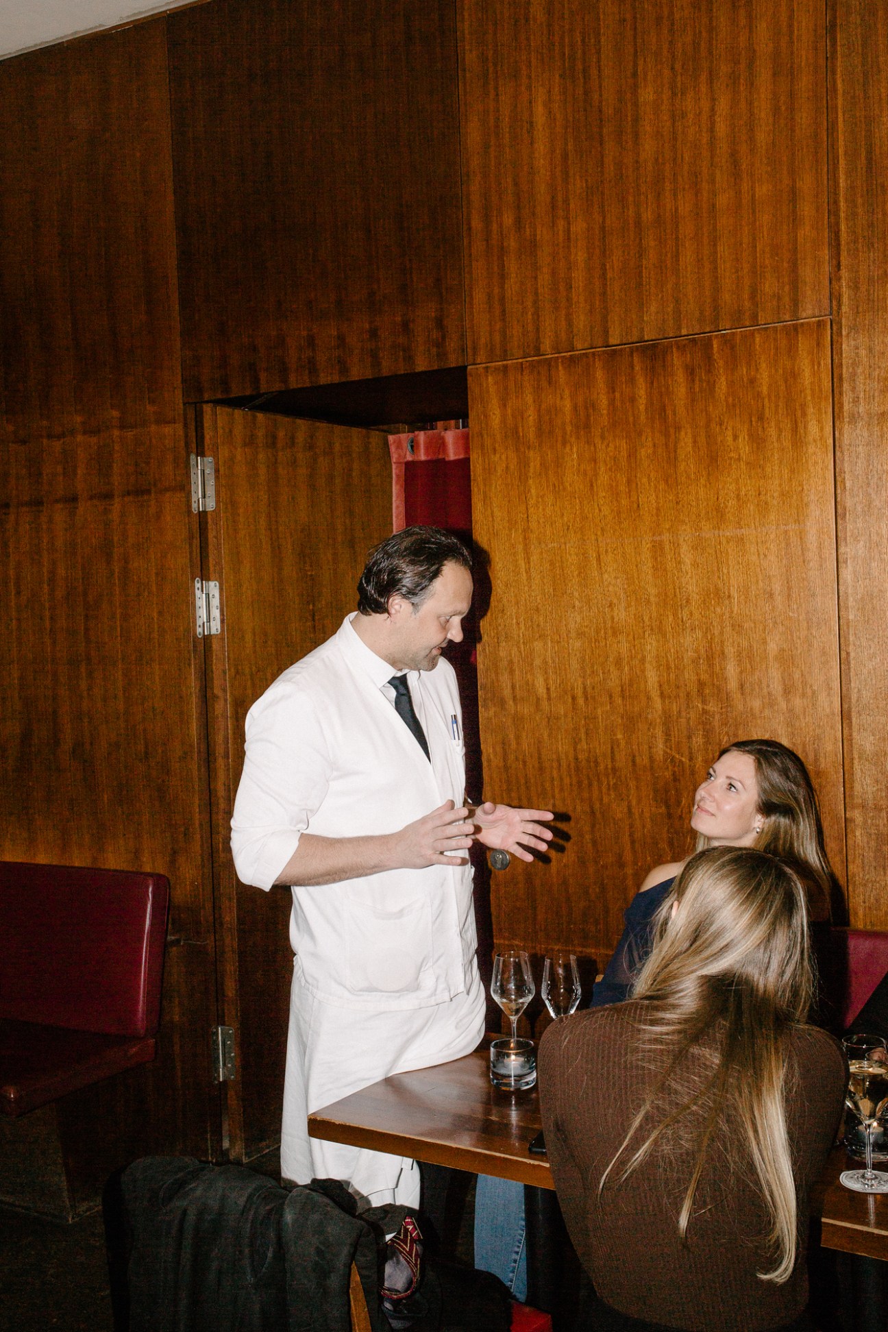 A waiter in white coat speaks with two blonde women seated at a table in an upscale restaurant
