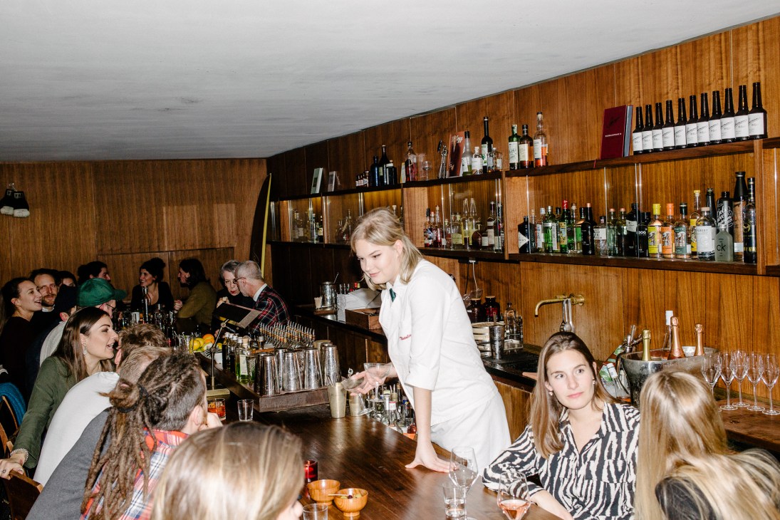 A blonde bartender in a white jacket serves drinks behind a wood-paneled bar with bottles on shelves