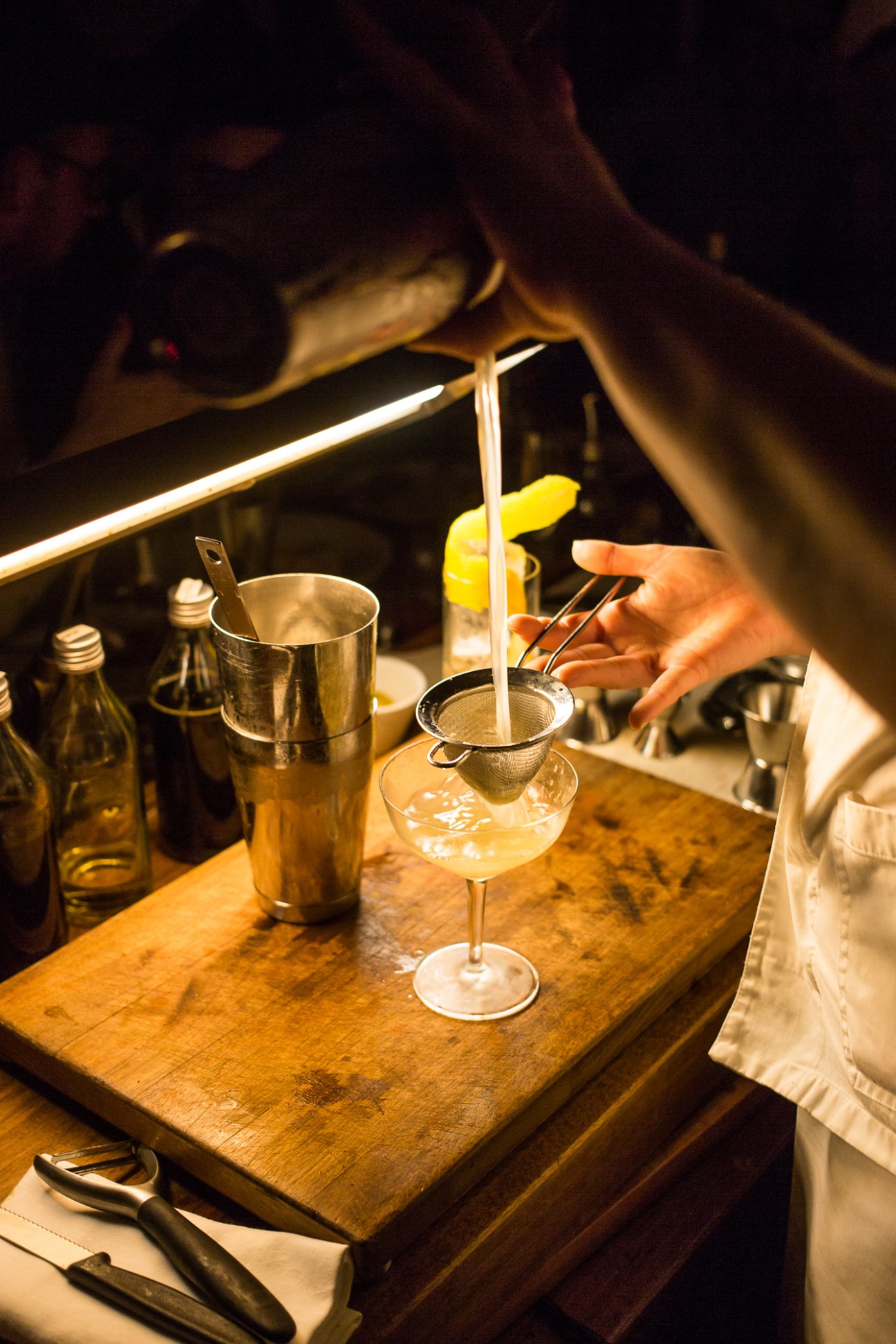 Bartender straining cocktail through fine mesh strainer into coupe glass at dimly lit bar
