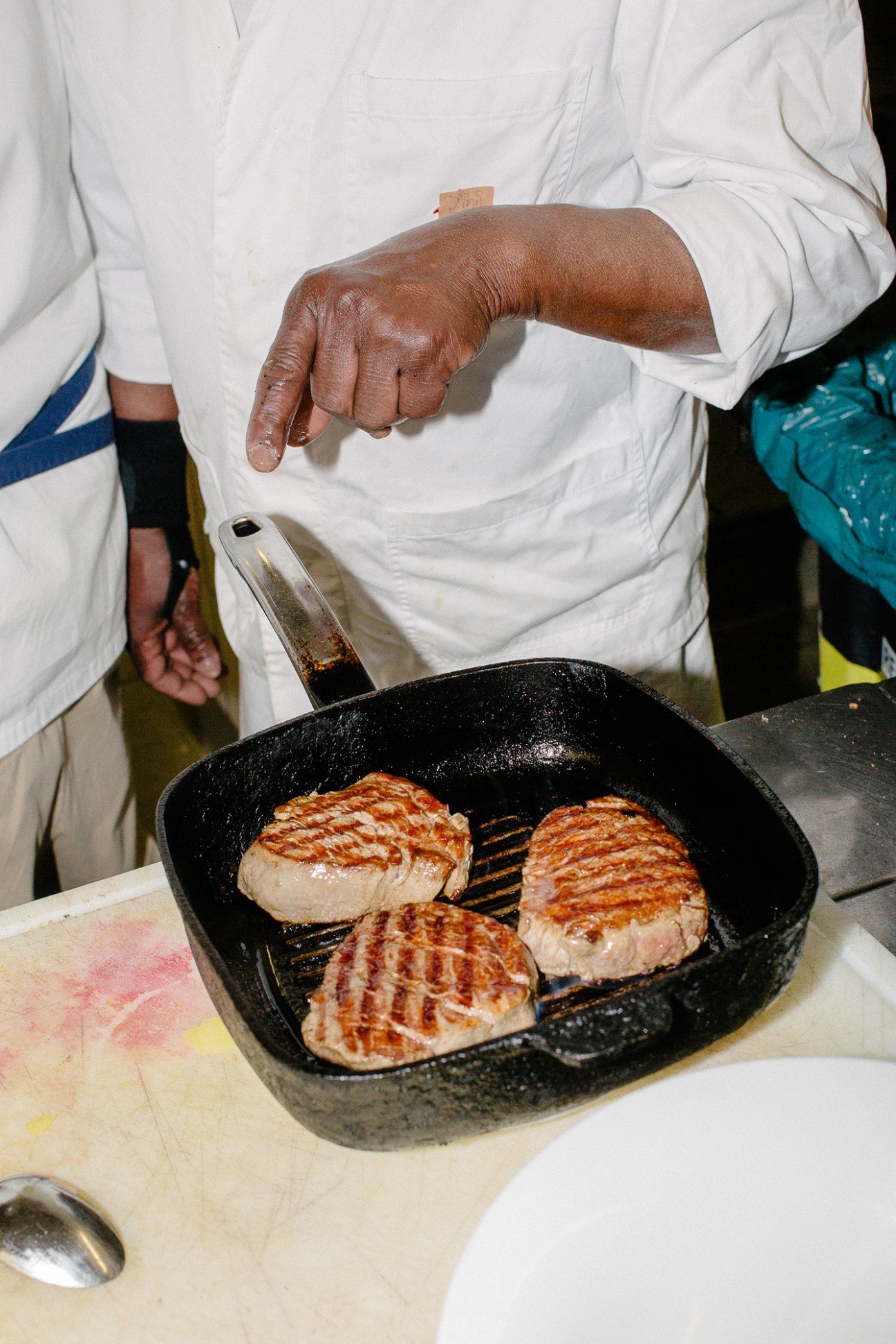 Chef in white coat grilling three pieces of meat in a black grill pan