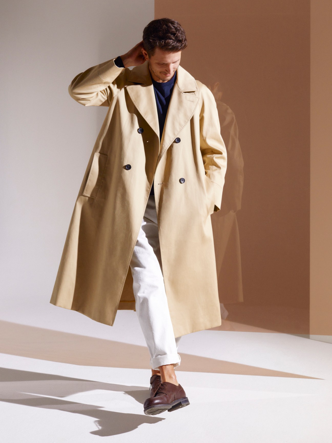 Man wearing beige double-breasted trench coat with navy sweater, white trousers and brown leather shoes