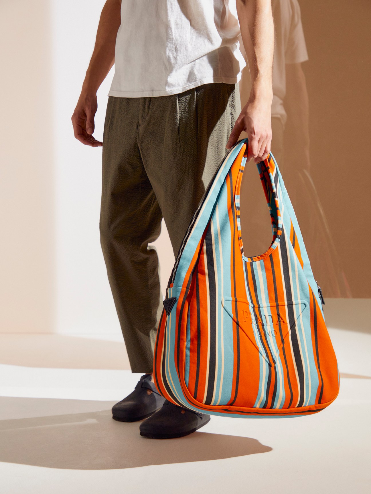 Man wearing white t-shirt and dark trousers holding colorful striped canvas beach tote bag with Birkenstock sandals