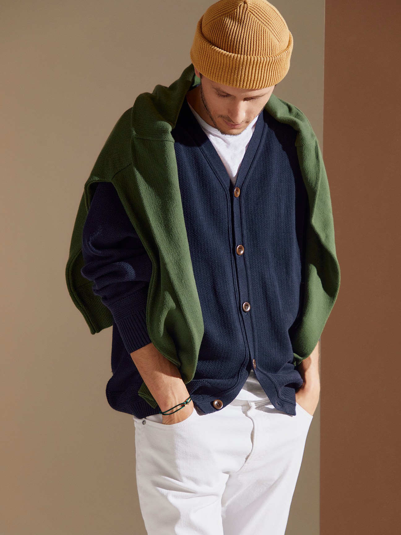 Man wearing mustard beanie, green cardigan over navy cardigan with white trousers