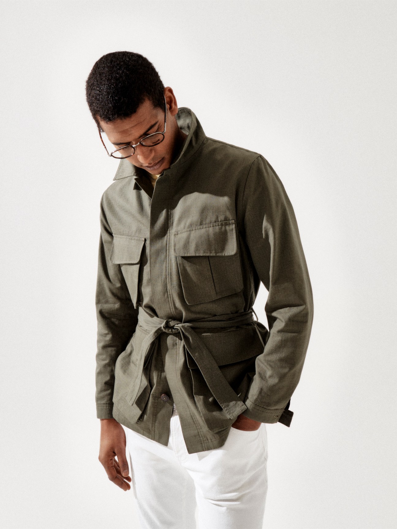 Model wearing olive green belted utility jacket with chest pockets, white trousers and glasses