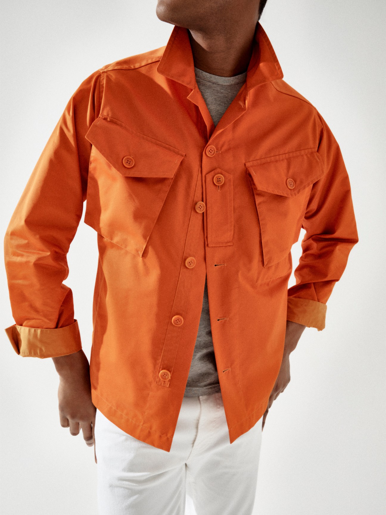 Model wearing orange button-up shirt jacket over grey t-shirt with white trousers