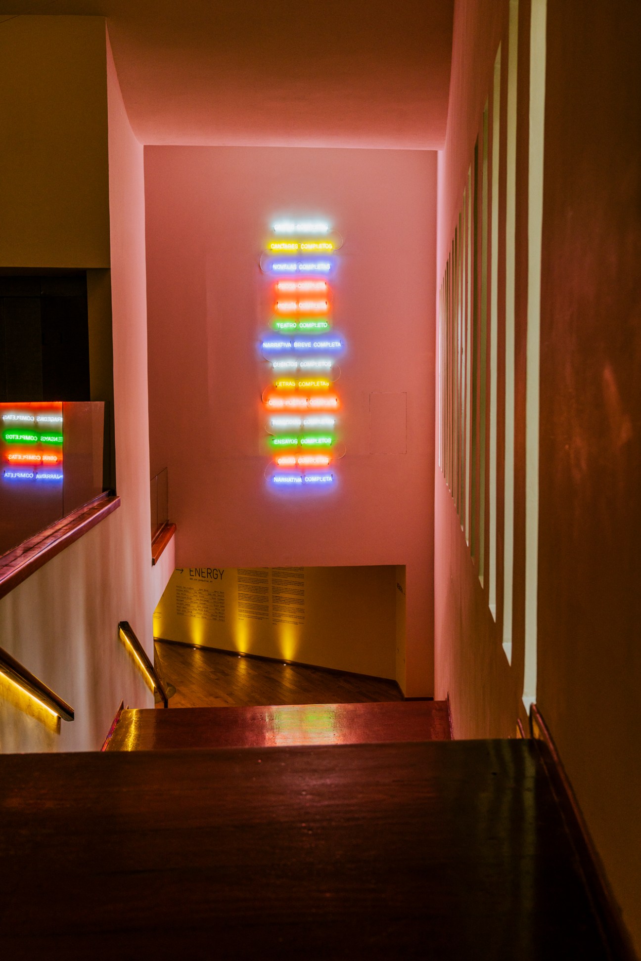 Neon light installation with colorful text stacked vertically in a gallery stairwell.