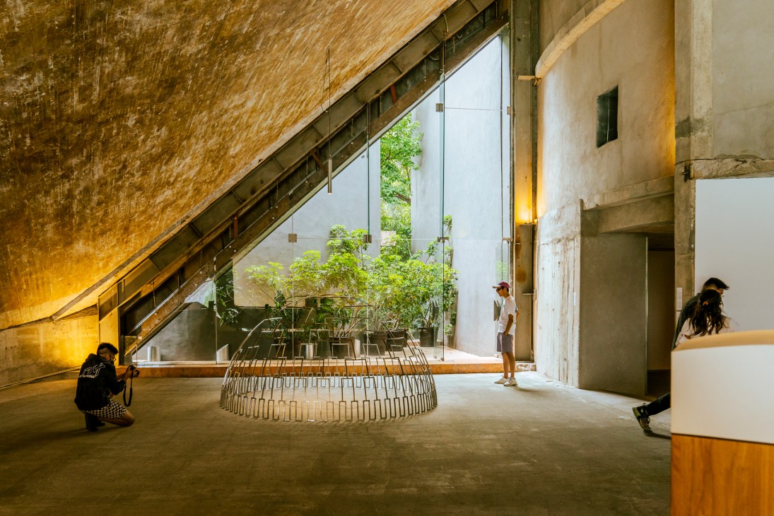 Visitors photograph an art installation in a modernist gallery space with dramatic angled ceiling and large windows overlooking a garden.