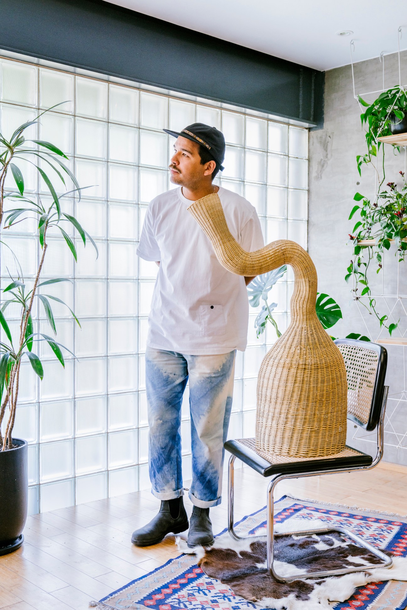 Man in white shirt standing next to woven chair in bright interior space with plants