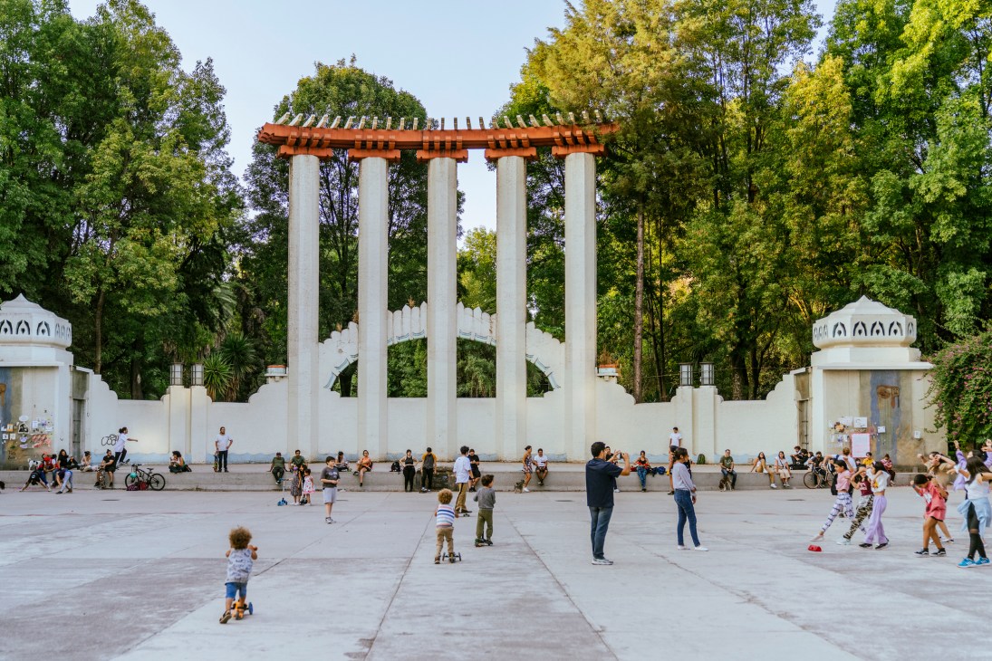 White monument with classical columns and decorative elements in a public square surrounded by trees