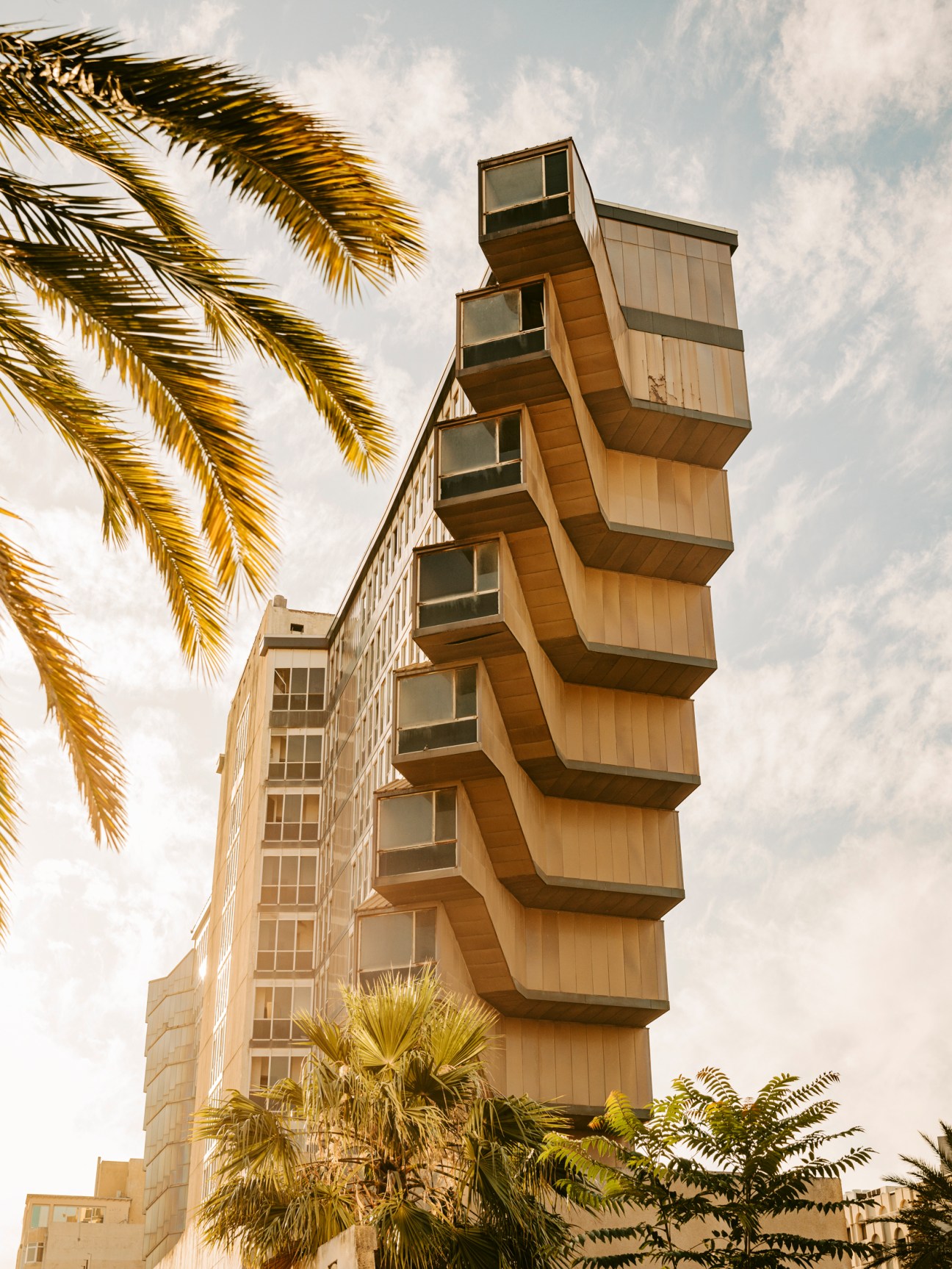 Modernist building with stacked hexagonal balconies jutting from facade, surrounded by palm trees against cloudy sky