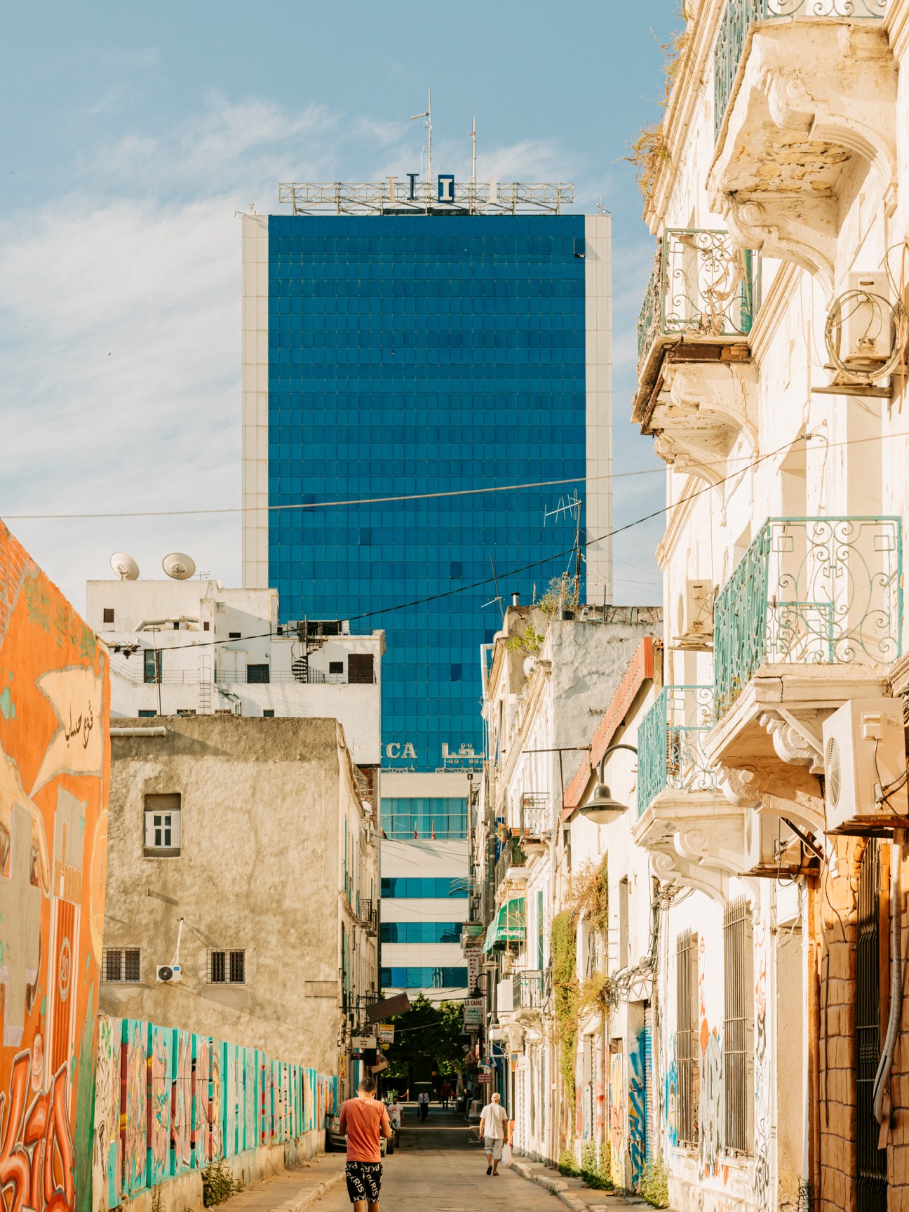 A narrow street in Tunis with colonial architecture, ornate balconies, and a modern glass tower in the background