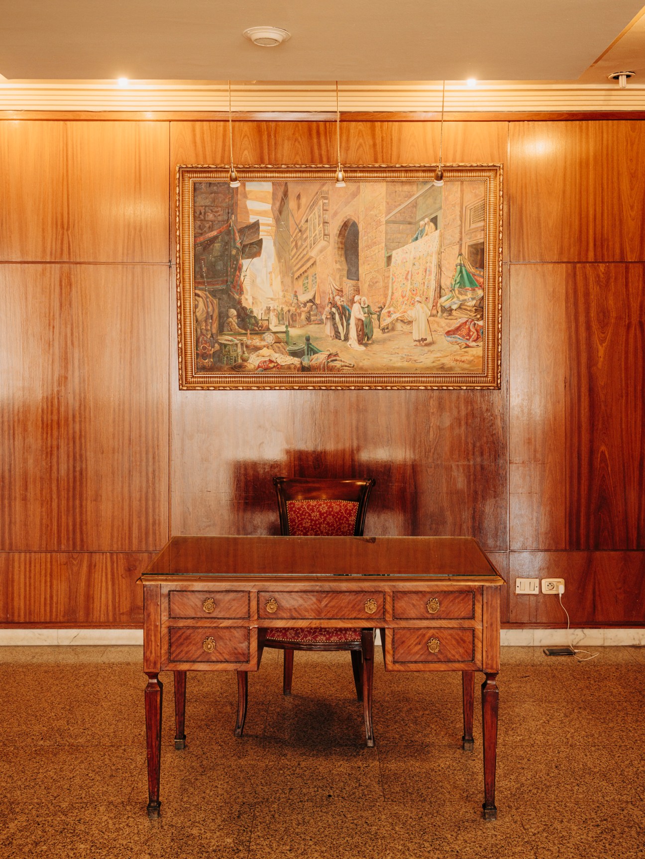 Wood-paneled office interior with ornate desk, upholstered chair, and framed Middle Eastern marketplace painting