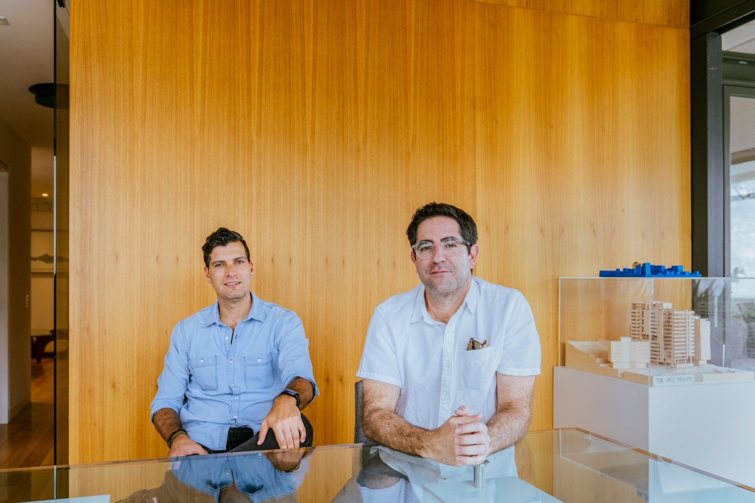 Two men in light blue shirts sitting at a glass table in a modern office with wood paneling and architectural models