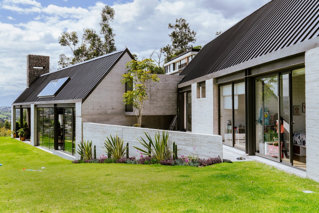 Modern residential building with angular metal roofing, concrete walls, and large glass windows surrounded by landscaping