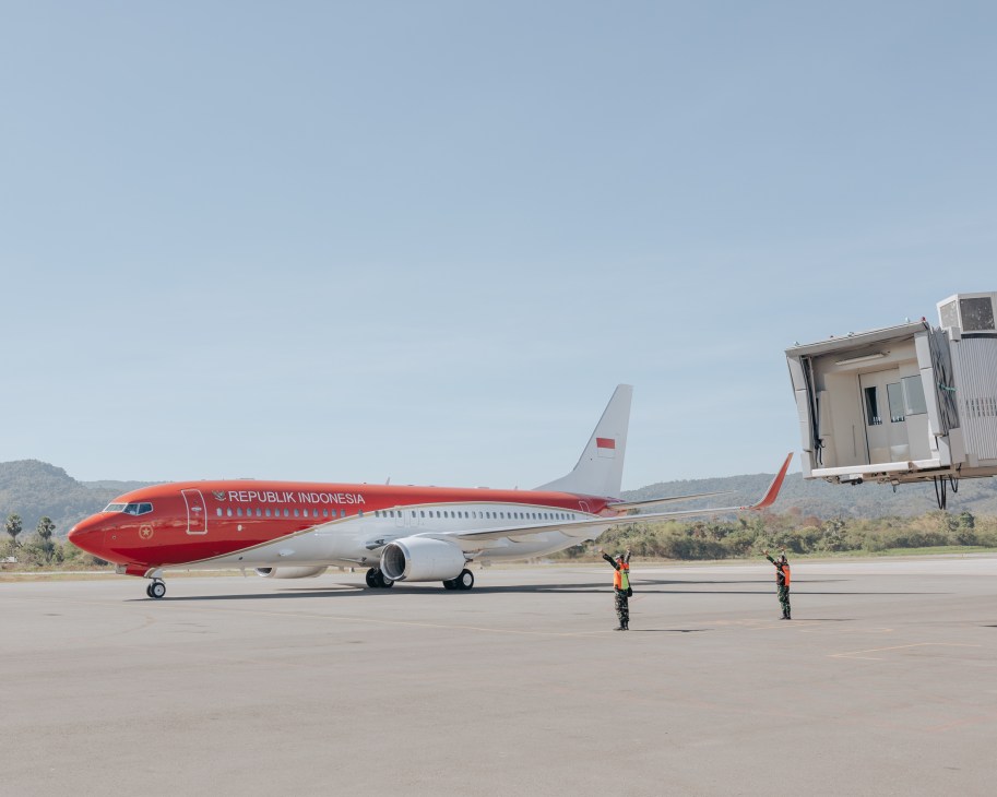 Red and white Indonesian presidential aircraft on tarmac with ground crew and terminal building in background