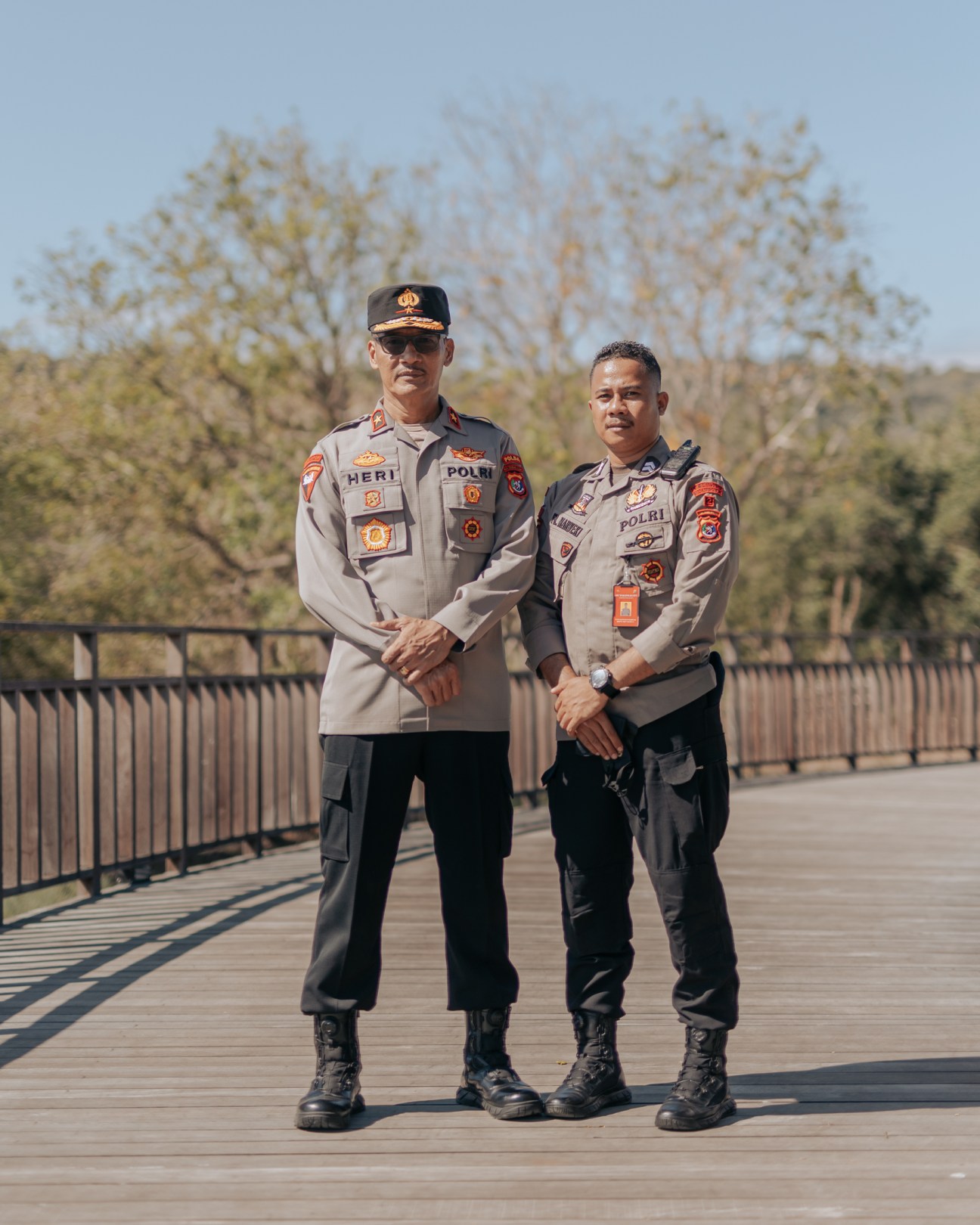 Two Indonesian police officers in tan uniforms stand on wooden boardwalk with metal railings and trees in background