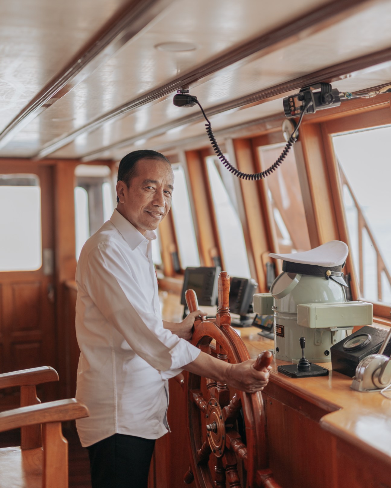 Indonesian President Joko Widodo standing at the wooden steering wheel of a boat's bridge, smiling at camera