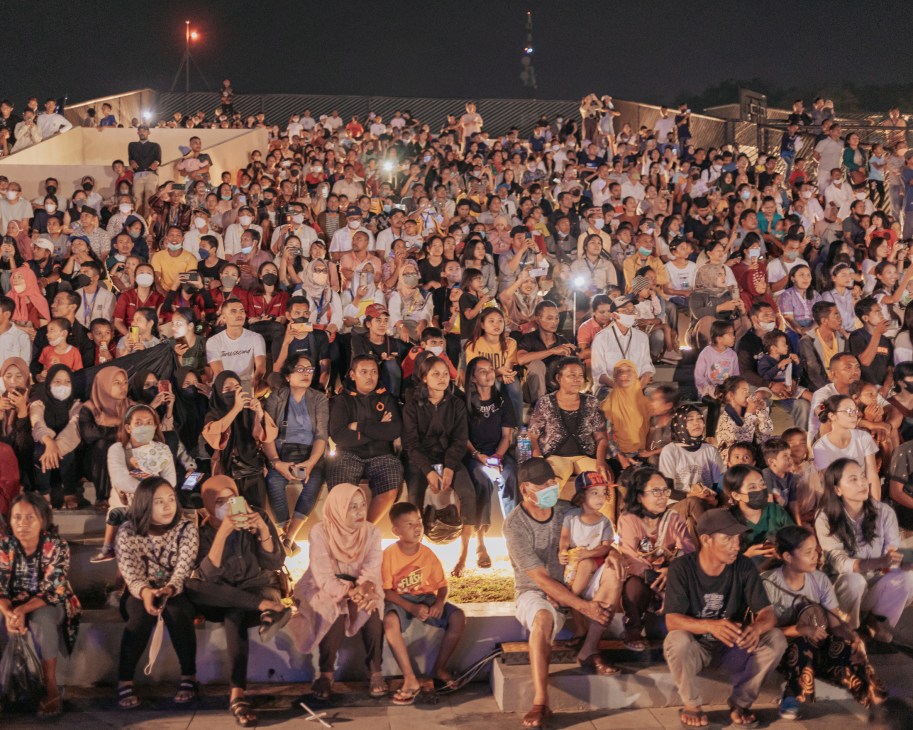 Large crowd of people seated in tiered outdoor venue at night, many wearing masks and holding phones