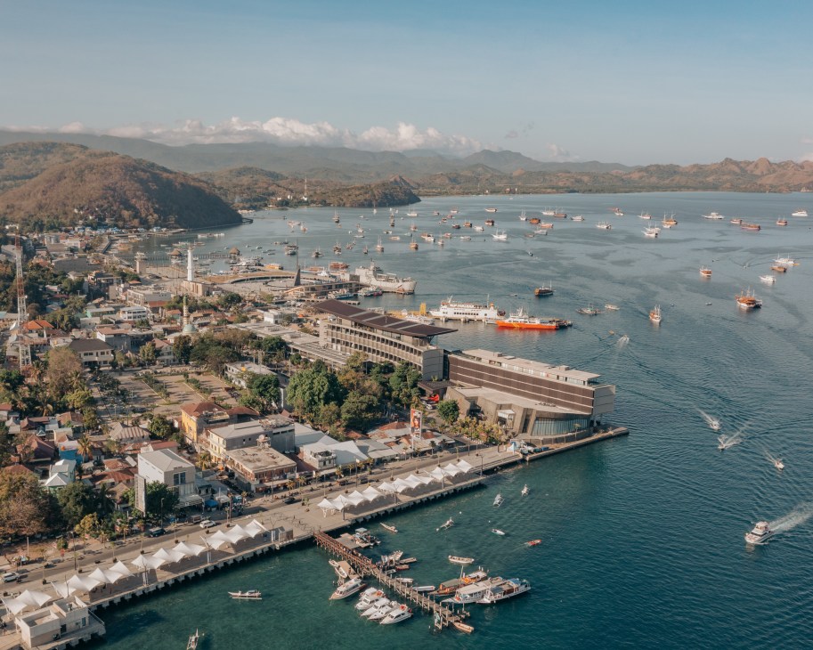 Aerial view of Labuan Bajo's waterfront with modern buildings, ferry terminal, and numerous boats anchored in the bay