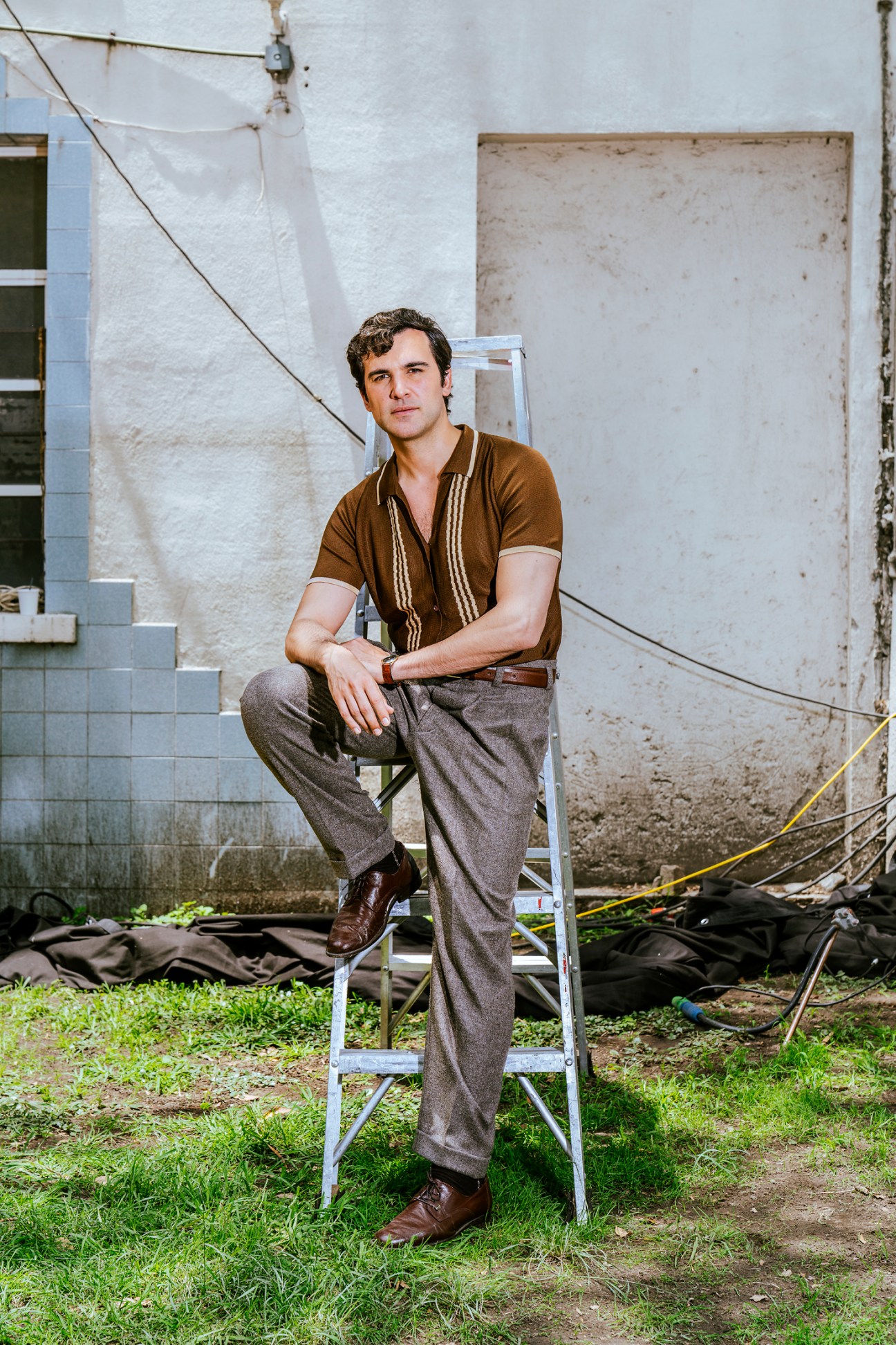 Juan Pablo di Pace sitting on a ladder on a film set, wearing a brown shirt and gray pants