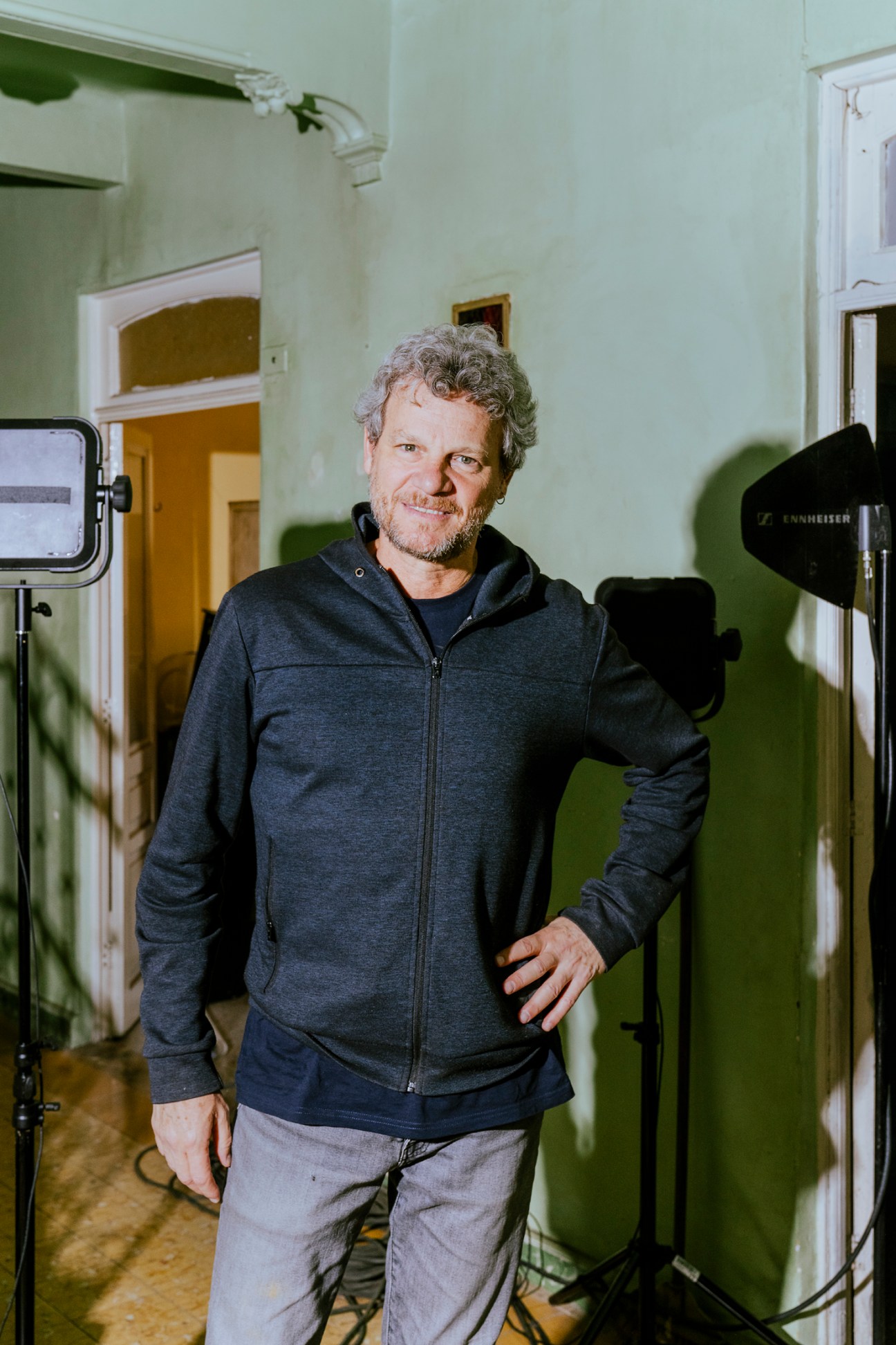 Middle-aged man with curly gray hair wearing dark zip-up hoodie standing among film equipment in white-walled interior