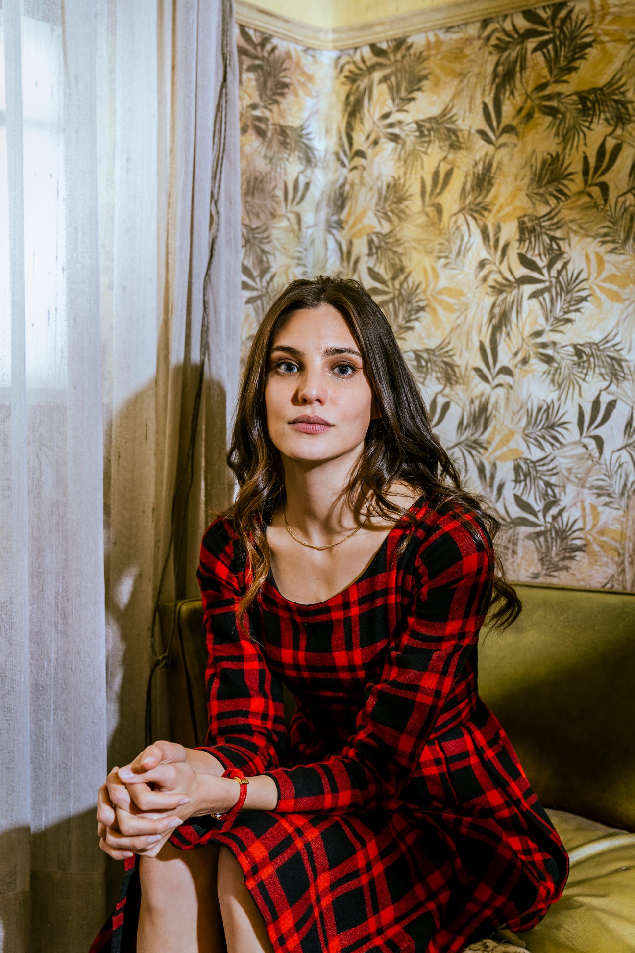 Argentinian actor Macarena Achaga in red plaid dress sitting on yellow furniture in ornate room with palm wallpaper