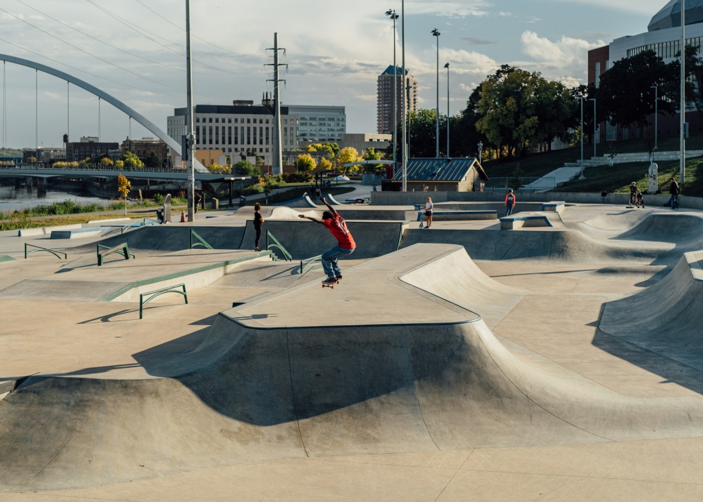 Skateboarders practice tricks at Lauridsen Skatepark in Des Moines with city skyline and river in background.
